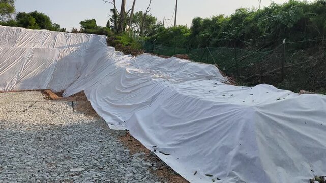 A newly cut soil slope is covered with temporary plastic sheeting. This tarp prevents rainwater erosion to ensure site safety. Workers will remove the cover once grass planting begins.