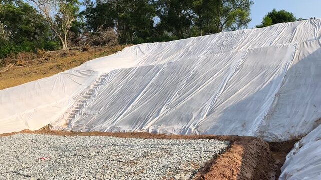 A newly cut soil slope is covered with temporary plastic sheeting. This tarp prevents rainwater erosion to ensure site safety. Workers will remove the cover once grass planting begins.