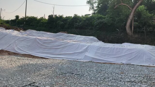A newly cut soil slope is covered with temporary plastic sheeting. This tarp prevents rainwater erosion to ensure site safety. Workers will remove the cover once grass planting begins.