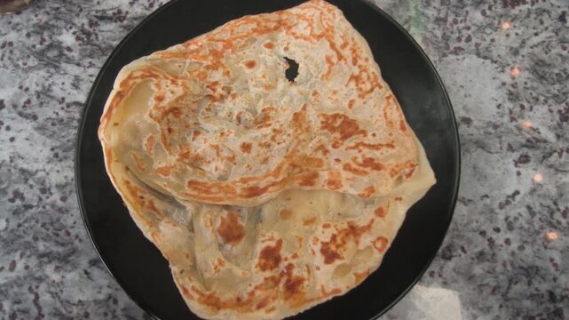 A piece of classic roti canai is served on a plate with savory dhal curry. This represents the staple, basic preparation of the dish in Malaysia. It remains immensely popular for breakfast.