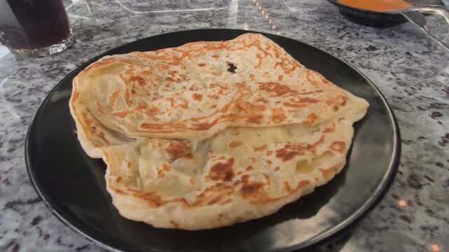A piece of classic roti canai is served on a plate with savory dhal curry. This represents the staple, basic preparation of the dish in Malaysia. It remains immensely popular for breakfast.