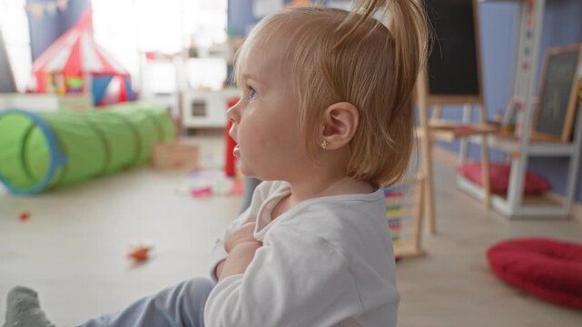 Toddler girl sitting on a green ottoman in a playroom building with socks visible and a ponytail, hands resting; joyful curiosity.