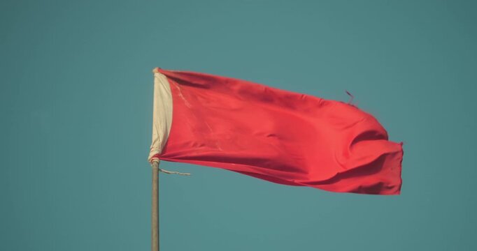 Red flag flying strongly in the wind against a clear blue sky, signaling danger, caution, warning, or a significant event unfolding
