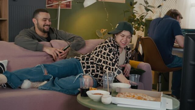 Young Caucasian man working on laptop and arguing with noisy flatmates, watching TV, laughing aloud and relaxing on couch in shared house. Clip is ideal for student lifestyle or co-living service ads