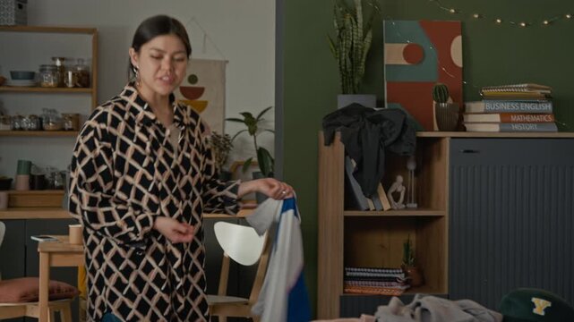 Frustrated young Asian woman throwing messy clothes at lazy young Caucasian and HIspanic roommates lying on sofa in cluttered shared apartment. Youth gen Z lifestyle or roommate conflict commercials