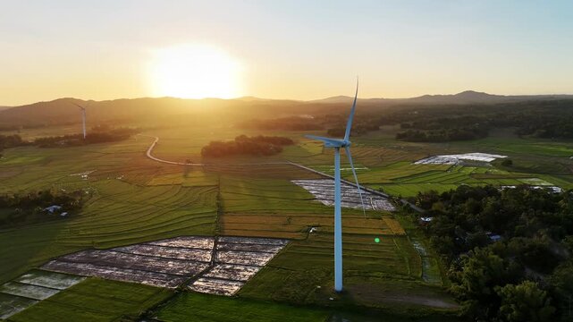 Aerial drone view of a white wind turbine and a second turbine in the distance rising above flat green and fallow rice paddies at sunset, San Lorenzo Wind Farm, Philippines