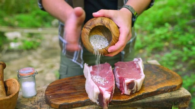 A person seasoning raw beef steaks with coarse sea salt from a wooden bowl while preparing a meal outdoors in a natural forest setting - Landscape Format.