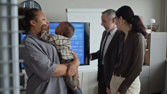 African American mother holding baby in office while Caucasian colleagues discuss business data on screen. Ideal for promoting family-friendly workplace policies and work-life balance