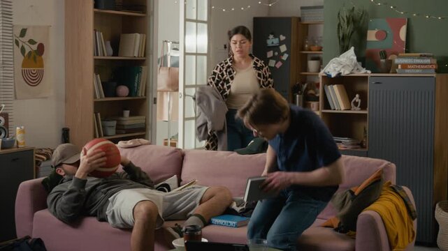 Young adult Asian woman throwing messy clothes at lazy young Hispanic man lying on sofa in shared dorm room, arguing and shouting. Student gen Z lifestyle and roommate conflict commercials