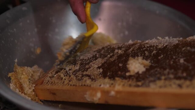 Close-up of a hand using a yellow spatula to scrape a brown crumbly mixture from a wooden board into a large metal bowl under warm indoor light