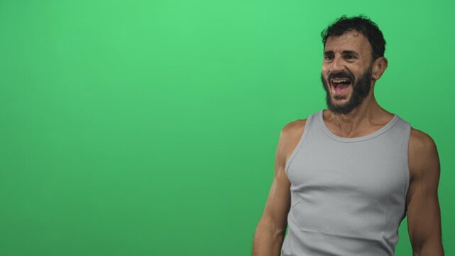 Man shouting with hands raised and bare arms against a studio green screen wearing a tank top and beard; joy.