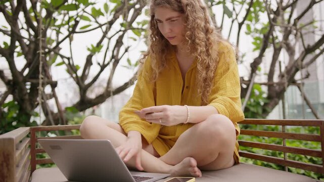 Curly haired young woman works online using her laptop and smartphone on an outdoor daybed during the bright daytime hours in the balcony of the hotel