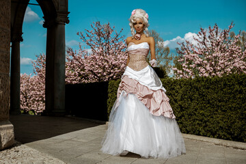 Naklejka premium Gorgeous aristocratic woman in an ornate pink and white baroque ballgown and powdered wig posing in a vibrant spring garden with blooming cherry trees and roses