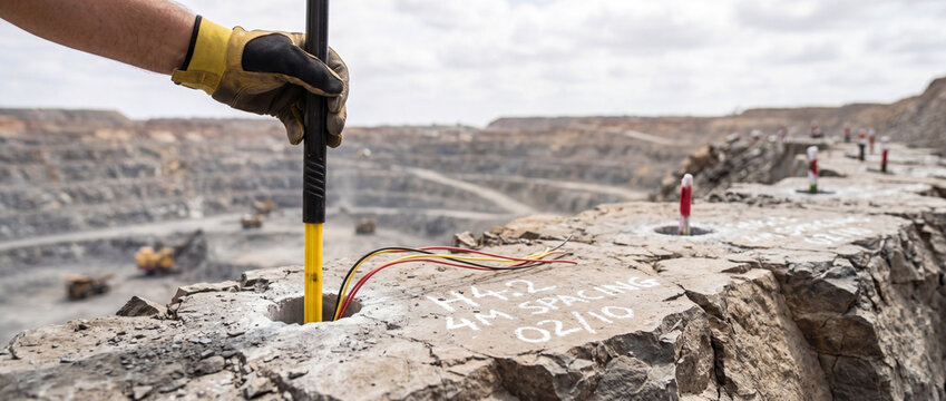 Mining worker inserts a drill rod and connects explosive wires in marked blast holes along the rim of an open-pit quarry, preparing for a controlled rock blasting operation.