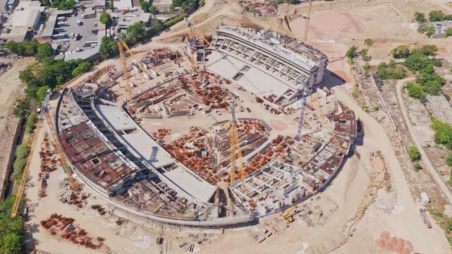 High aerial perspective showing the ongoing construction of a large, modern sports stadium or arena, with tower cranes working on the concrete and steel structure of the building