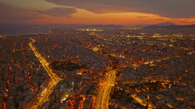 Stunning aerial perspective of thessaloniki, greece, at twilight, showing the illuminated city streets, bustling traffic, and a beautiful orange sky as evening descends over the urban landscape