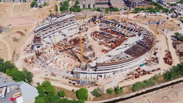 Majestic aerial view showing the construction of a large circular stadium, highlighting the intricate network of cranes, concrete structures, and reinforcement bars shaping the future arena