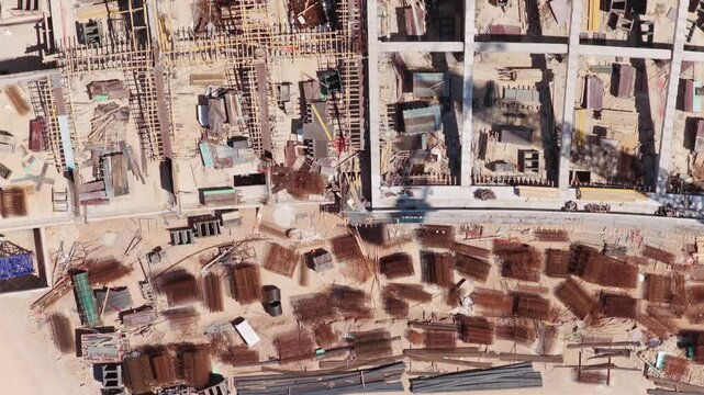 Aerial top-down perspective following a construction site, showing the building foundation and various materials like scaffolding, rebar cages, and steel beams scattered on the ground