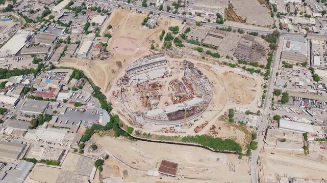 High aerial view showing the massive construction site of a modern circular stadium or arena, highlighting the developing infrastructure and urban growth in the surrounding industrial area