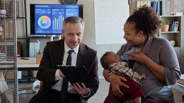 African American woman feeding baby with bottle while discussing document with Caucasian colleague in office. Clip ideal for HR and family-friendly workplace campaigns