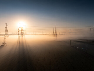 Fototapeta premium Power transmission towers in morning fog with sunlight creating a serene industrial landscape