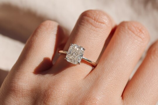 Minimalist close-up of hand wearing sparkling lab-grown emerald or cushion  cut  diamond engagement ring, soft natural light on smooth neutral background. Ethical bridal jewelry, sustainable luxury.