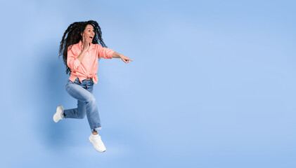 Obraz premium A woman is jumping in mid-air while laughing and pointing to the side. She wears a light pink shirt and blue jeans. The background is a bright blue. She seems to have fun and energy
