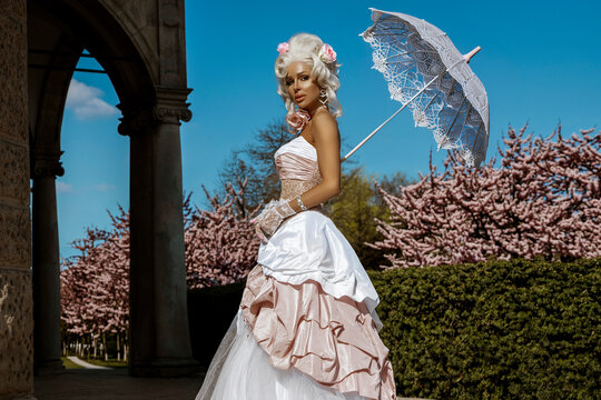 Elegant woman in a historical Rococo style pink and white gown holding a lace parasol in a beautiful blooming spring garden