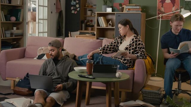 Diverse young adult students studying in shared apartment, Caucasian female showing frustration while flatmate watching noisy video on laptop. Student lifestyle and exam stress commercials