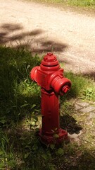 Czerwony hydrant nadziemny stoi stabilnie na trawiastym poboczu tuż obok piaszczystej ścieżki. Obiekt rzuca wyraźny cień w pełnym letnim słońcu, otoczony naturalną, zieloną roślinnością. © tomo81