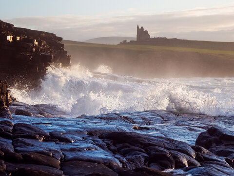 Sunlight glints off wet, rounded stones along the shoreline. Waves crash against the rocks with force and rhythm. Castle Classiebawn in the background. County Sligo, Ireland. Travel and tourism.