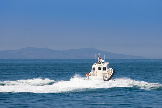 Rear view of a white coast guard patrol boat cruising at high speed on the blue sea water, leaving a white foam wake with mountains in the background.