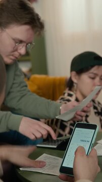 Vertical of multiethnic young male and female zoomers calculating household utility bills using smartphone calculator app while sharing accommodation. Student budgeting app, personal finance service