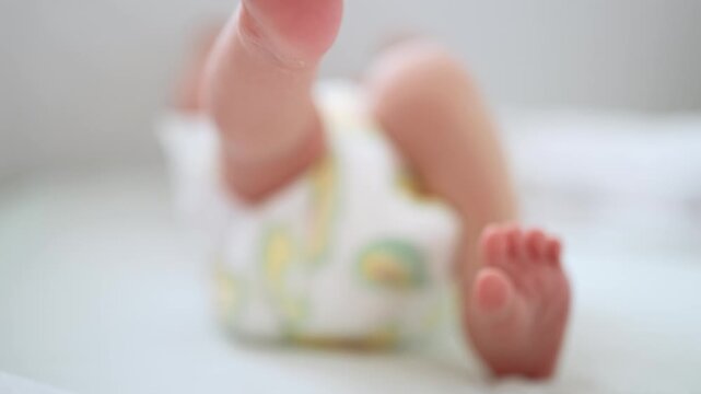 Close up of a playful newborn infant kicking tiny legs and feet while lying on a changing table as a mother's gentle hands carefully change the baby's diaper in a bright room