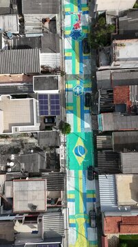 Vista a&eacute;rea de rua decorada com pinturas inspiradas na Sele&ccedil;&atilde;o Brasileira para a Copa, destacando tradi&ccedil;&atilde;o popular e manifesta&ccedil;&otilde;es culturais dos moradores.