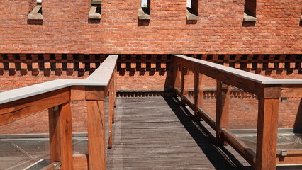 Drewniany pomost z balustradą prowadzi wzdłuż potężnego muru obronnego z czerwonej cegły. W górnej części budowli widoczne są regularnie rozmieszczone, wąskie otwory strzelnicze. © tomo81