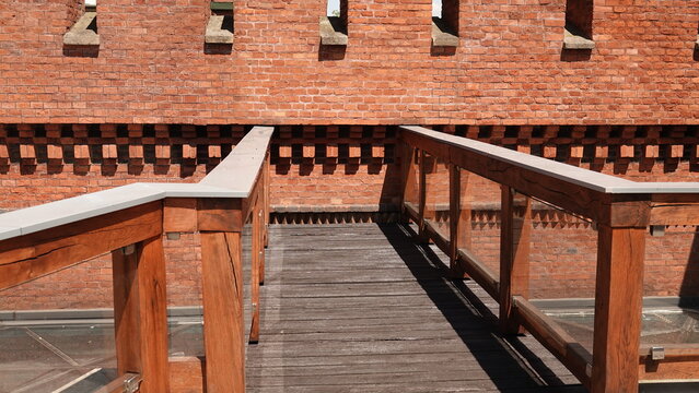 Drewniany pomost z balustradą prowadzi wzdłuż potężnego muru obronnego z czerwonej cegły. W g&oacute;rnej części budowli widoczne są regularnie rozmieszczone, wąskie otwory strzelnicze.