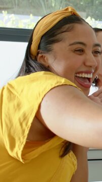 Vertical video: Laughing woman in yellow top reacting to friend, leaning in kitchen reaching arm