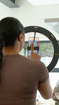 Vertical video: Tapping braided host framing phone in LED light in kitchen, cohost in apron mixing