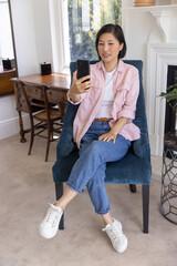 Obraz premium Asian woman in late 20s sitting in blue chair holding and viewing smartphone at home desk