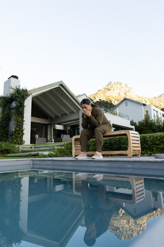 Curved wooden bench sitting at edge of still pool, covered patio seating and mountain reflecting