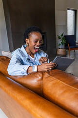 Obraz premium African American adult woman leaning over brown sofa, holding tablet, wearing smartwatch at home