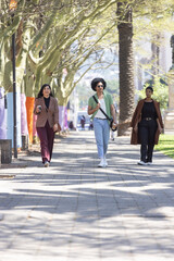 Three friends walking down tiled promenade under canopy, carrying coffee cup and crossbody bag © wavebreak3