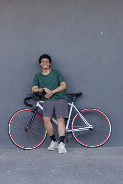 Man in 20s leaning against gray wall, holding white road bike with red rims, wearing wristwatch