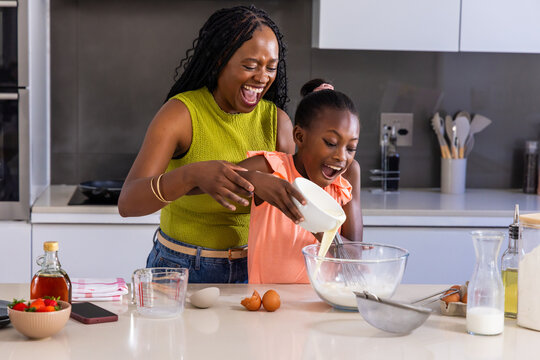 Mother and teenage daughter pouring batter into clear mixing bowl at kitchen island, whisk nearby