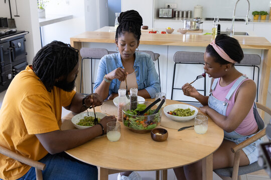 African American family eating green pasta at table in kitchen with teen daughter sharing salad