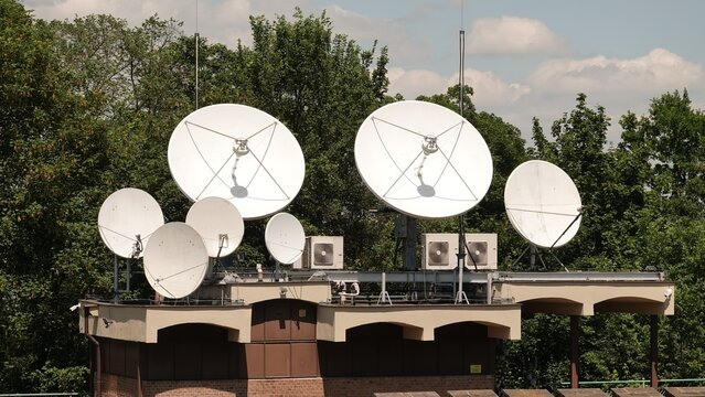Grupa białych anten satelitarnych o r&oacute;żnych rozmiarach zainstalowana na dachu budynku obok urządzeń klimatyzacyjnych. W tle widoczna jest gęsta ściana zielonych drzew w słoneczny dzień.