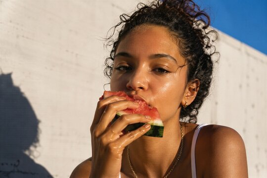 Hispanic woman eating fresh watermelon slice outdoors. Summer lifestyle portrait of girl enjoying juicy fruit in sunlight. Healthy food, refreshment, vacation, youth and summer mood concept