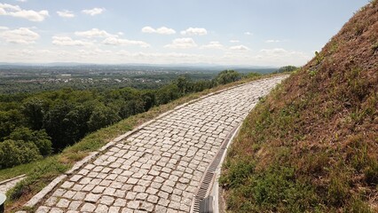 Kamienna, brukowana droga zakręca łagodnie wokół trawiastego zbocza wzniesienia. Z trasy roztacza się rozległa panorama na zielone lasy oraz nizinny horyzont pod częściowo zachmurzonym niebem. © tomo81