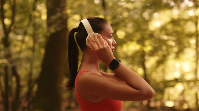 Woman wearing headphones and using smartwatch to browse songs and adjust volume before jogging, preparing for workout in nature, side view, 4K